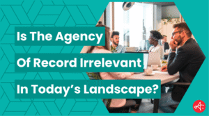 Team of marketing professionals engaged in a discussion around a conference table in a bright, modern office. Text overlay reads: Is The Agency of Record Irrelevant In Today’s Landscape?