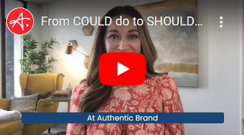 A woman with long brown hair wearing a floral red dress smiles while speaking to the camera. The background features a modern, well-lit office space with a yellow armchair, a plant, and abstract wall decor. A text overlay at the bottom reads, "At Authentic Brand."