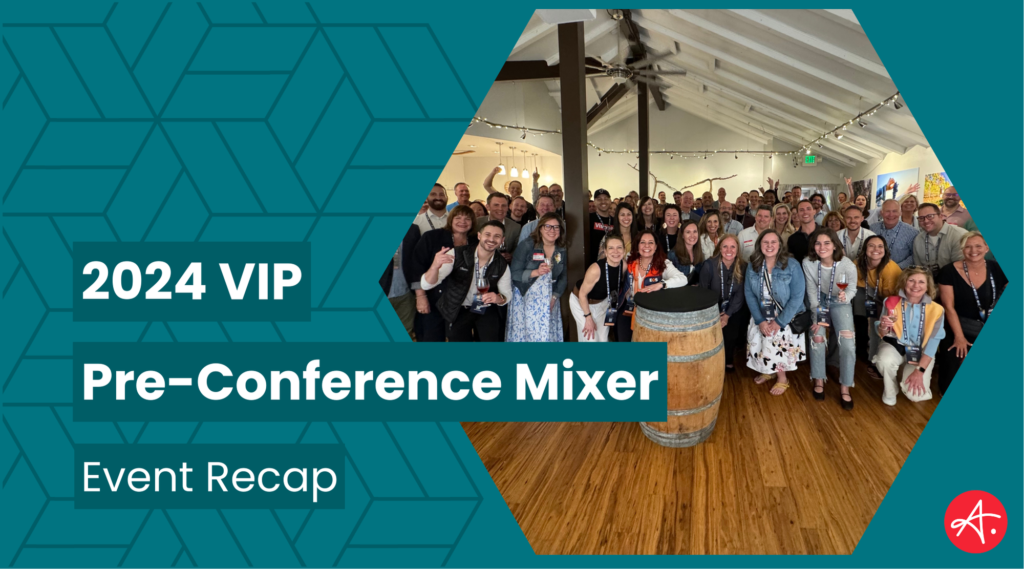 A large group of conference attendees posing together in a rustic venue with wooden floors and a central barrel display during the 2024 VIP Pre-Conference Mixer. The group is cheerful and diverse, enjoying the event atmosphere.