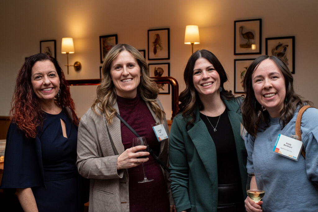 Four women stand side-by-side smiling for the camera at a social event, holding wine glasses, with framed artwork and wall sconces in the background.