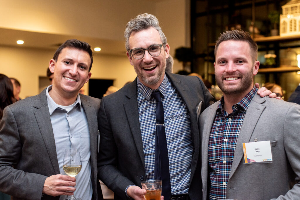 Three men dressed in suits pose cheerfully at a mixer, smiling at the camera while holding drinks.