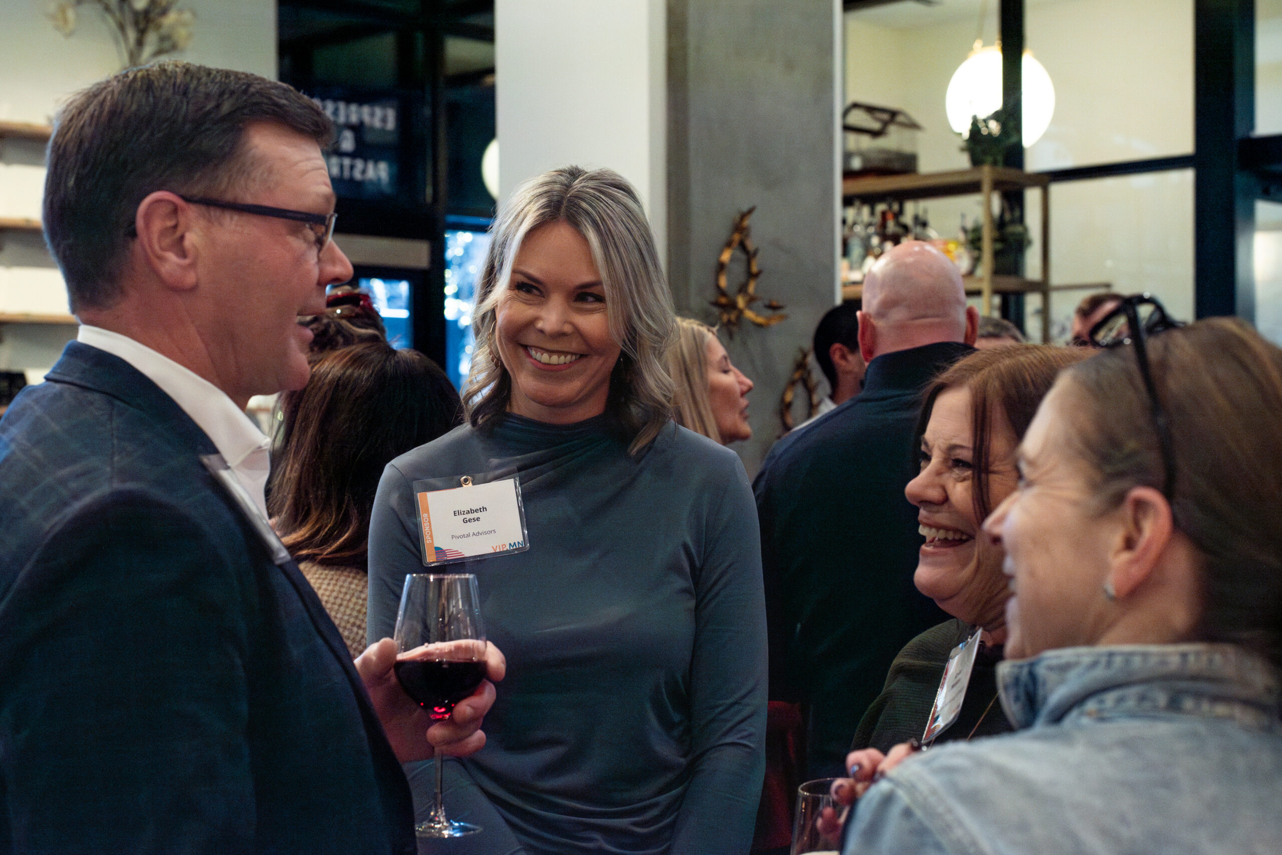 A group of professionals stands and chats warmly at a networking event; a woman in the center smiles while holding a glass of red wine.