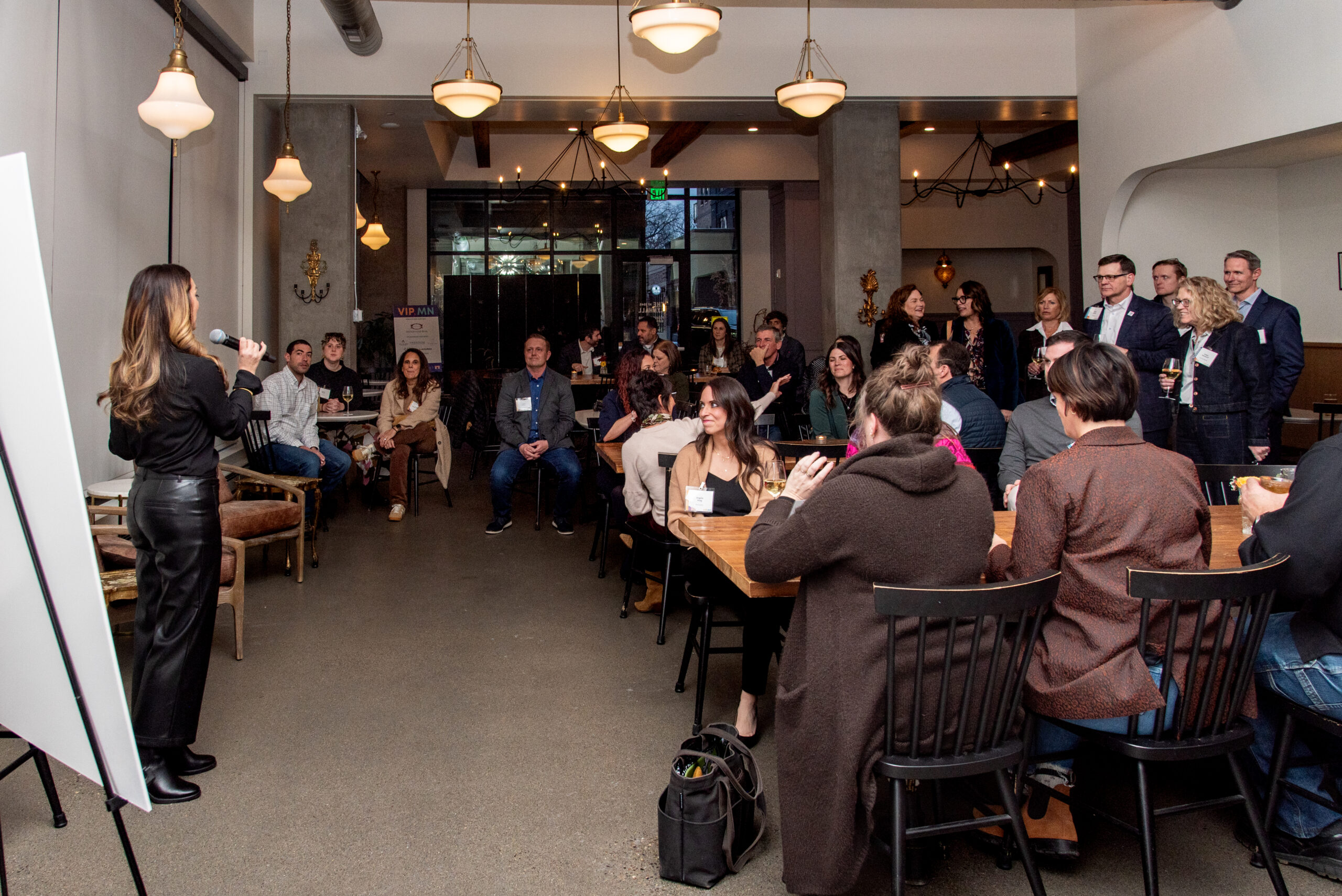 A woman holding a microphone addresses an audience seated at tables in a warmly lit event space.