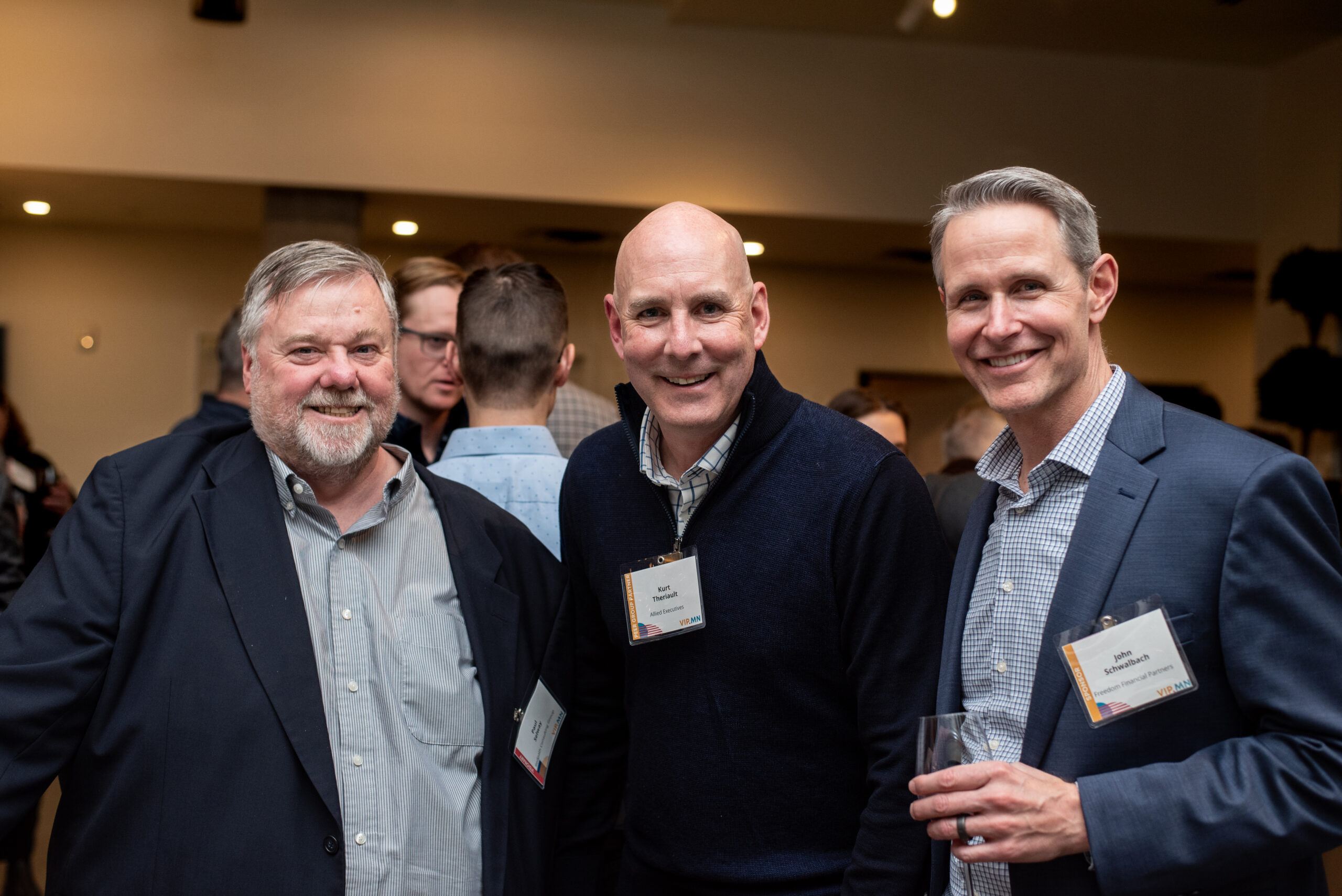 Three men smile for the camera at a professional mixer, holding drinks and wearing business attire with name tags.