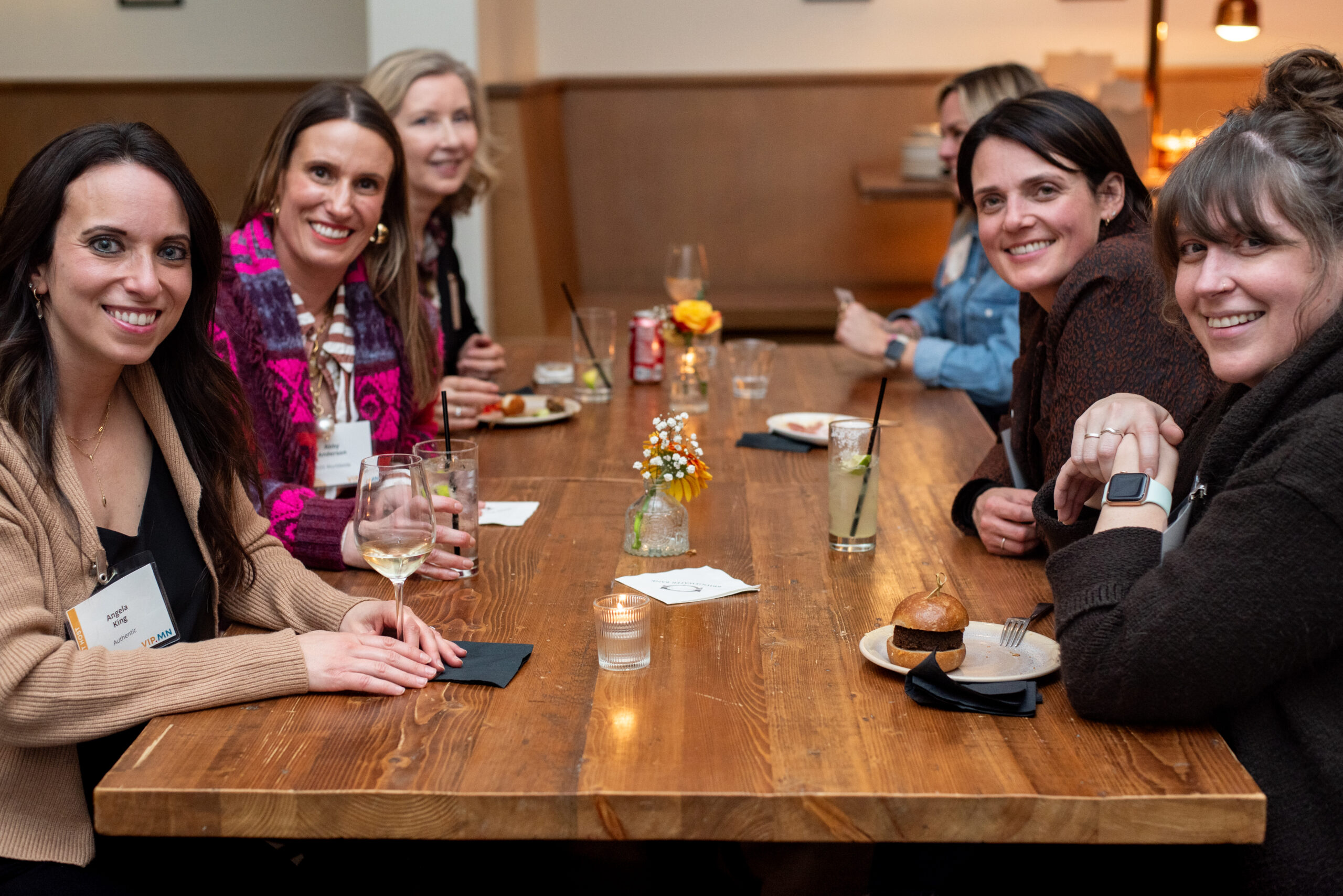 A group of women sits around a long wooden table, smiling and enjoying drinks and food at a networking mixer.