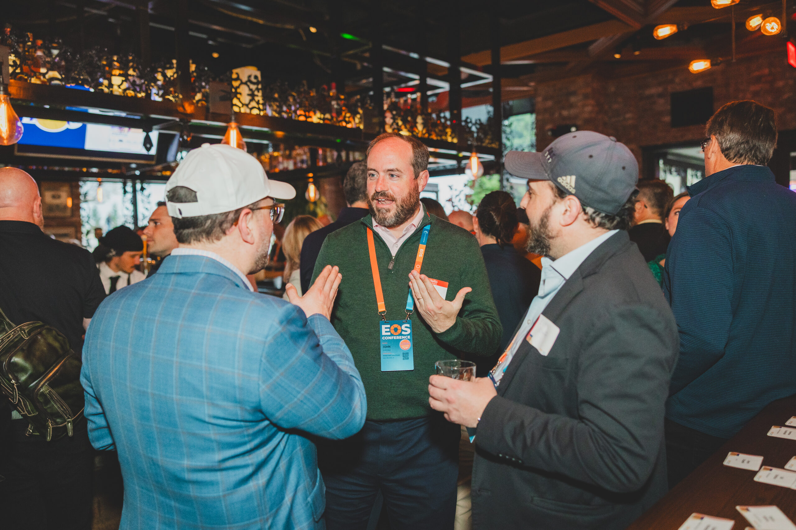 A man in a green sweater speaks animatedly to two other attendees wearing EOS badges while holding a drink at a busy bar.