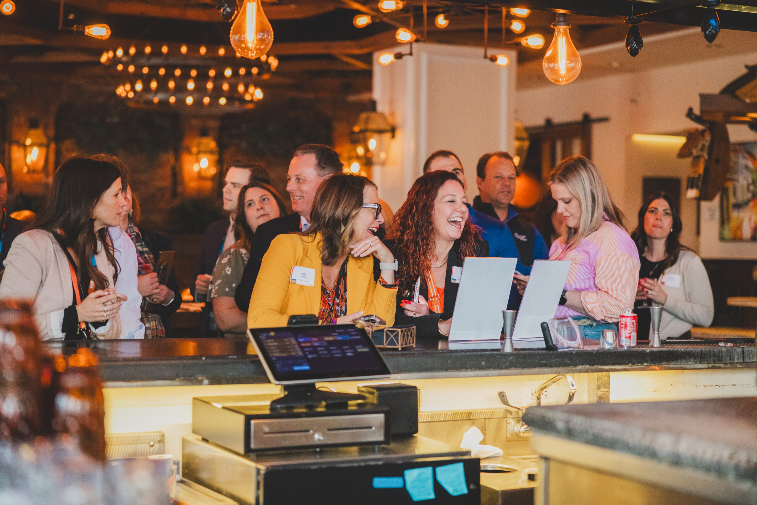 A crowd of attendees gathers at a bar counter, laughing and interacting while checking in or browsing materials near two laptops.
