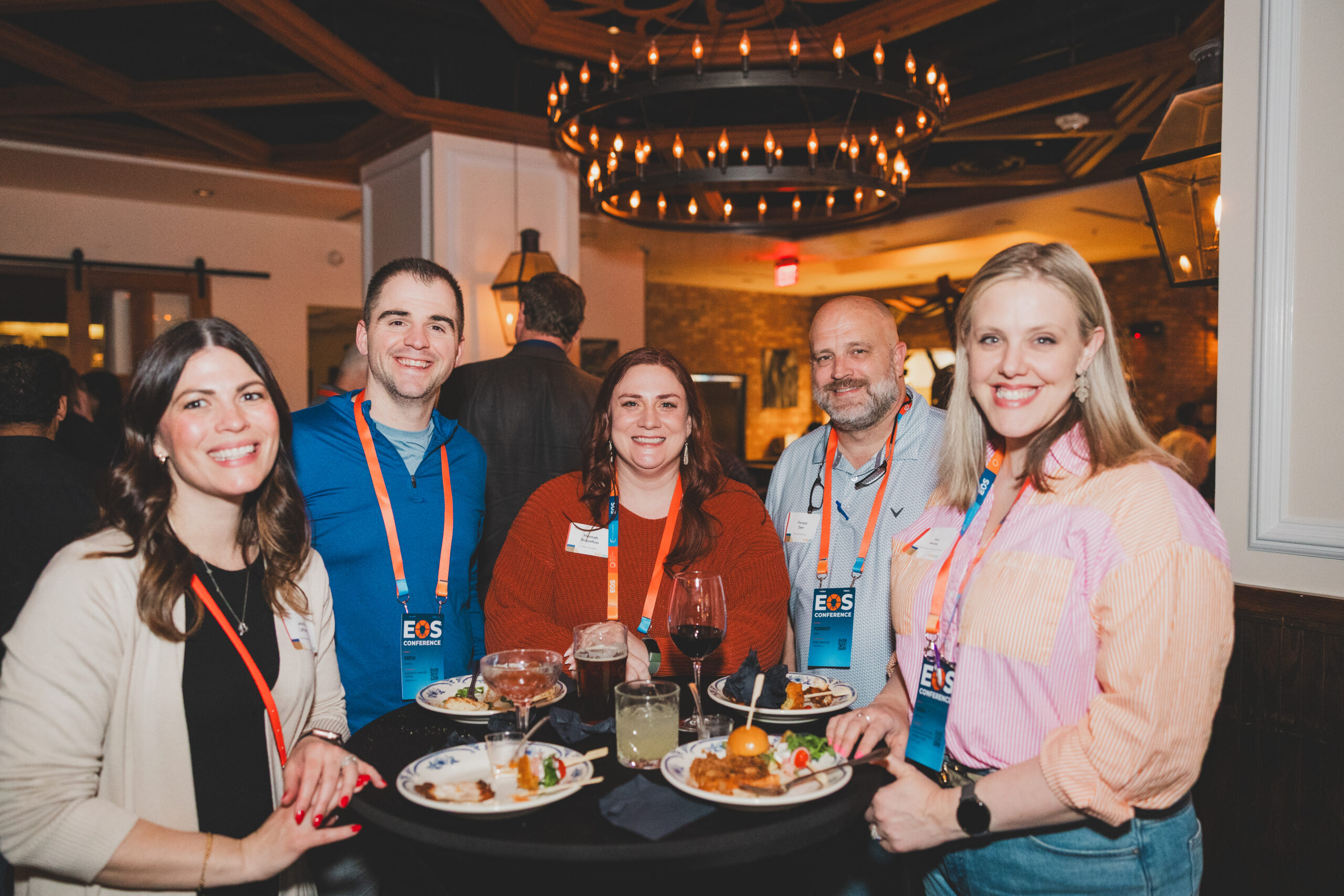 A cheerful group of five conference attendees—three women and two men—stand close together smiling, surrounded by warm lighting and brick walls.